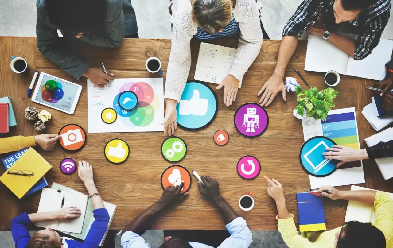 A diverse group collaborates around a wooden table, discussing ideas with colorful icons, notebooks, and drinks A diverse group collaborates around a wooden table, discussing ideas with colorful icons, notebooks, and drinks